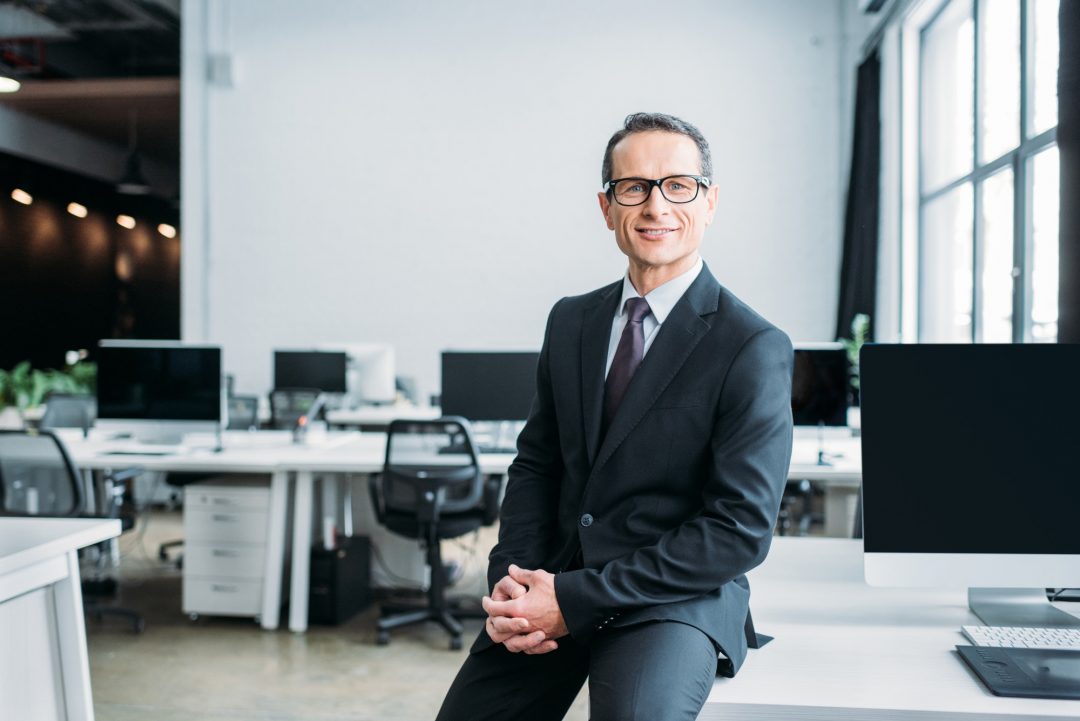 smiling businessman in eyeglasses sitting on table in office